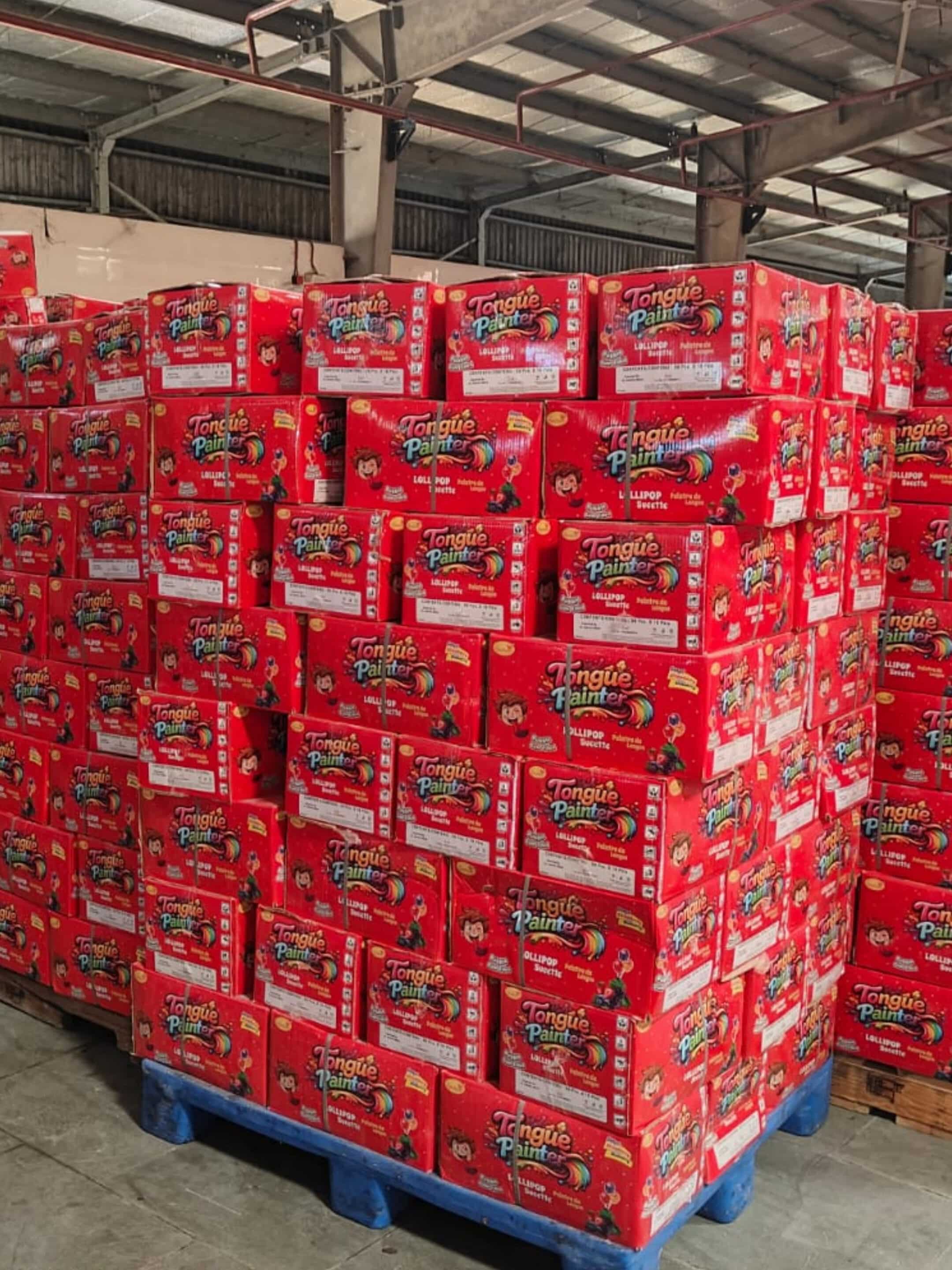 Stacks of red Tongue Painter toy boxes on blue pallets in a warehouse storage area. The boxes show cartoon characters and product branding.